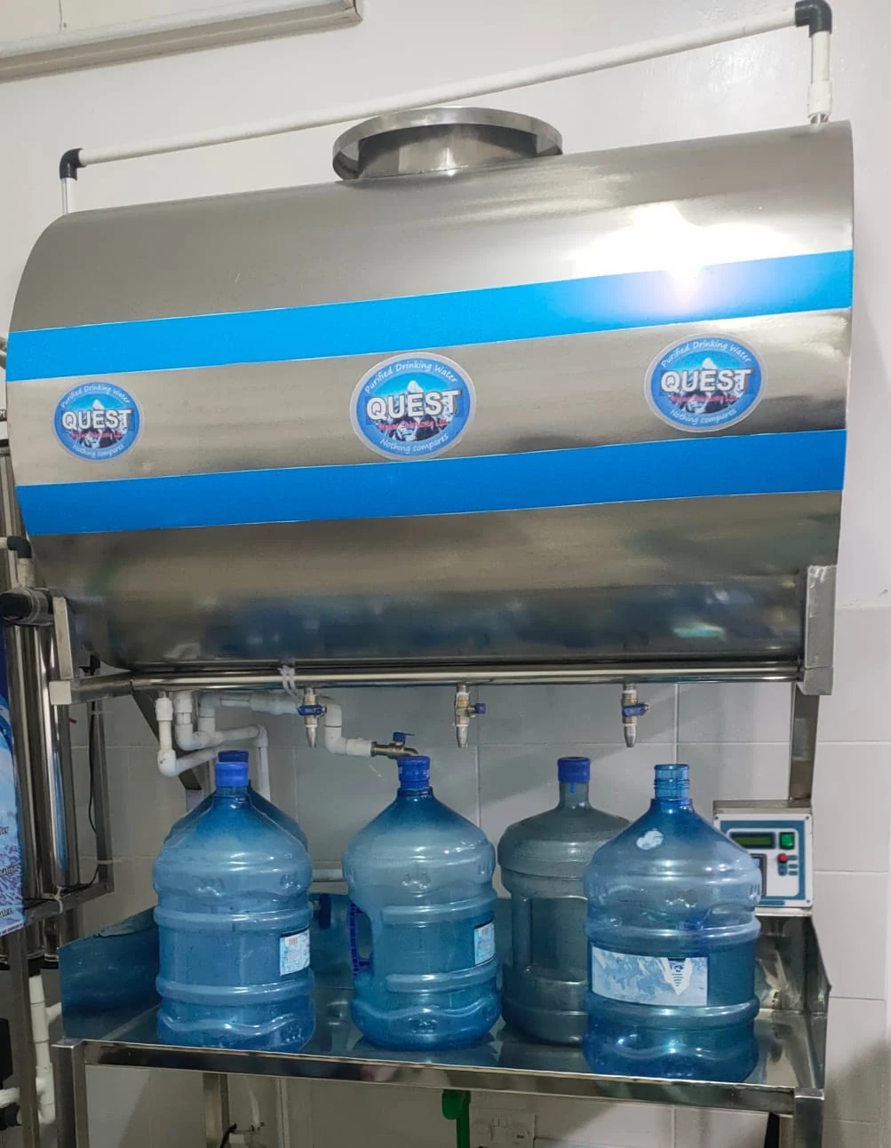 Stainless steel water vending tank with blue bands and four taps above refillable bottles in a refill shop